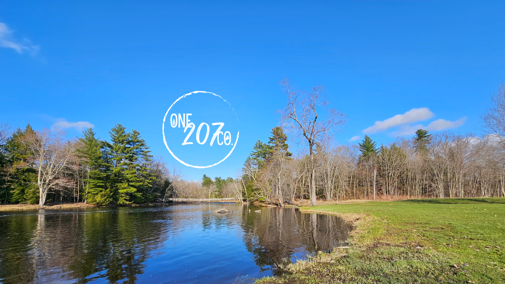 View of a stream winding north with green grass on the right and trees in the background with a beautiful blue sky. The One207Co logo appears above the stream.